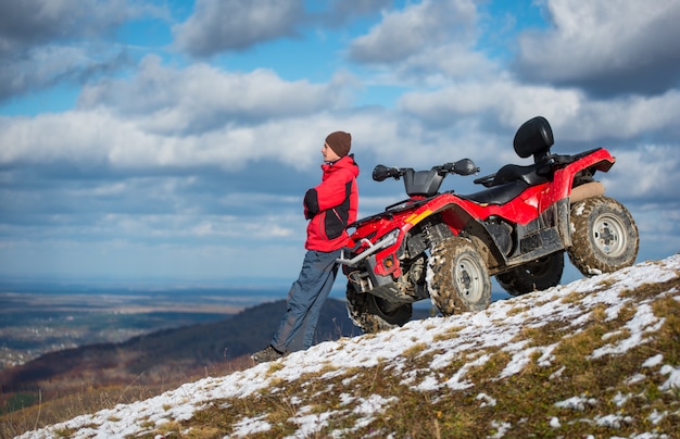 ATV moto perto do homem olha para a distância na encosta da montanha de neve na frente do céu azul nublado com espaço da cópia