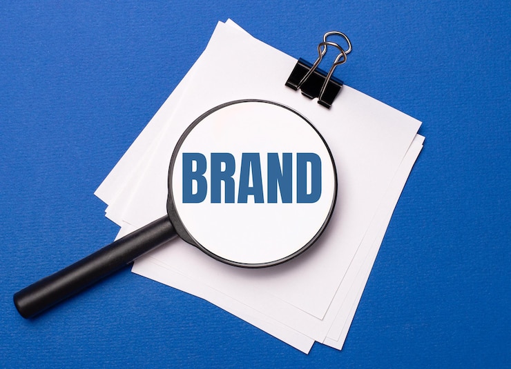 Auf blauem Grund weiße Blätter unter einer schwarzen Büroklammer und darauf eine Lupe mit dem Text BRAND