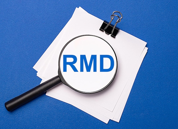 Auf blauem Hintergrund weiße Blätter unter einer schwarzen Büroklammer und darauf eine Lupe mit dem Text RMD Required Minimum Distributions