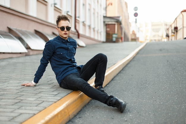 Auf dem Bürgersteig sitzt ein hübscher junger Mann in einem blauen Hemd, schwarzen Jeans und Turnschuhen