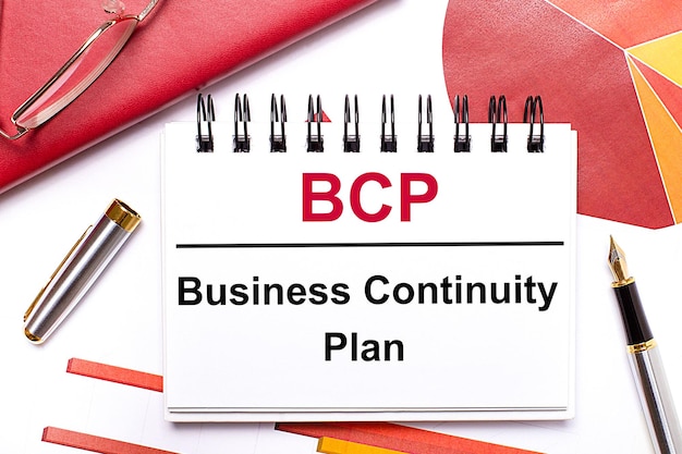 Auf dem Desktop befindet sich ein weißes Notizbuch mit dem Text BCP Business Continuity Plan, einem Stift in Burgund und roten Tischen und goldgerahmten Gläsern Geschäftskonzept