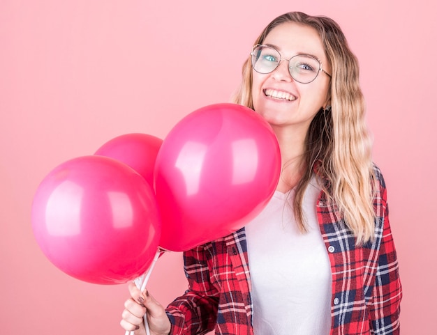 Auf dem Foto fühlt sich eine fröhliche dunkelhäutige Frau sehr glücklich und aufgeregt, hält Luftballons und lacht, verbringt ihre Freizeit auf einer Party, trägt ein schönes kariertes Hemd und eine Brille