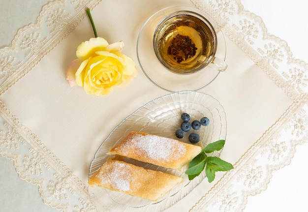 Auf dem Teller liegen zwei Scheiben Apfelkuchen, Heidelbeeren und Minzblätter, daneben eine Tasse Tee, und der Tisch ist mit einer lebenden Rose geschmückt