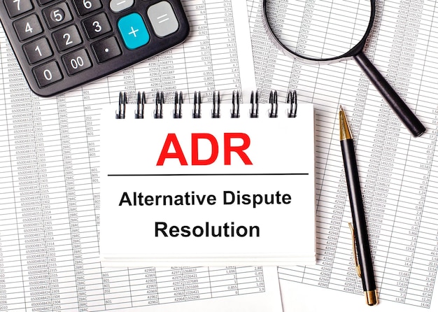 Auf dem Tisch liegen Berichte, eine Lupe, ein Taschenrechner, ein Stift und ein weißes Notizbuch mit dem Text ADR Alternative Dispute Resolution