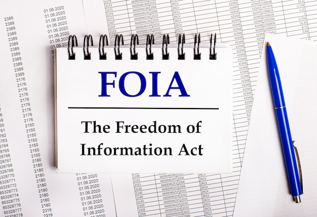 Auf dem Tisch liegen Diagramme und Berichte, auf denen ein blauer Stift und ein Notizbuch mit dem Wort FOIA The Freedom of Information Act liegen