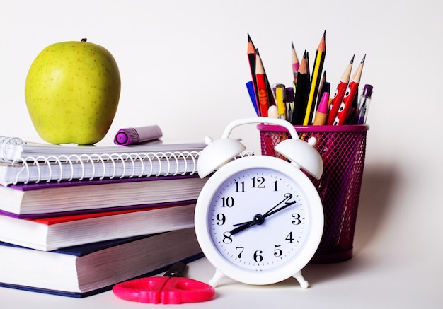 Auf dem Tisch liegen ein Stapel Bücher, Schreibwaren, ein weißer Wecker und ein Apfel