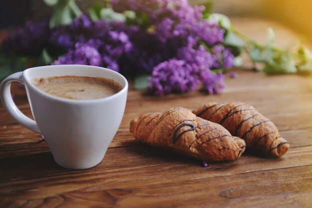 Auf dem Tisch stehen ruhige Croissants und eine Cappuccino-Tasse. Ein Blumenstrauß aus Flieder auf einem Holztisch.