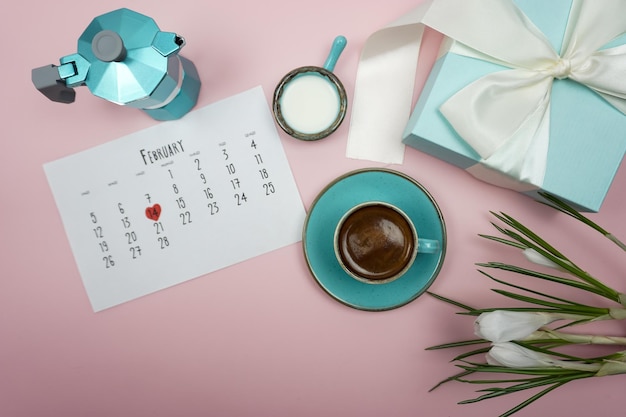 Auf dem Tisch steht eine Geysir-Kaffeemaschine, eine Tasse Kaffee, ein Kalender für Februar und Blumen