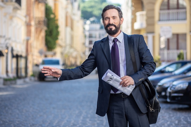 Auf dem Weg. Professioneller fröhlicher Geschäftsmann, der auf eine Fahrt wartet, während er eine Zeitung hält