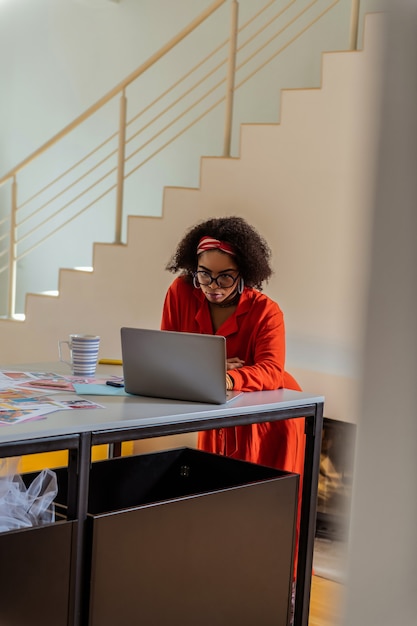 Auf den Bildschirm schauen. Beschäftigte ansprechende Mischlingsfrau, die Zeit bei der Arbeit verbringt, die seltsame Brille und rotes Outfit trägt