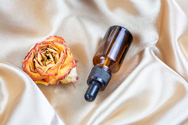 Auf den Falten aus Seidensatinstoff, umgeben von getrockneten Rosenblüten, liegt ein Behälter zur Aufbewahrung von Kosmetika aus dunklem Glas. Parfüm-, Serum- oder Lotionsflasche
