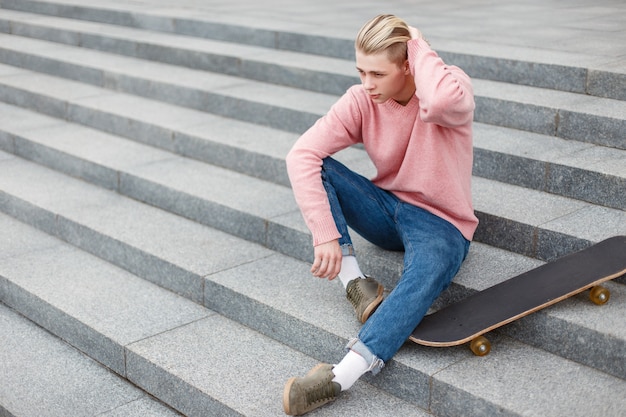 Auf den Stufen sitzt ein stilvoller gutaussehender Mann in modischer Kleidung mit einem Skateboard