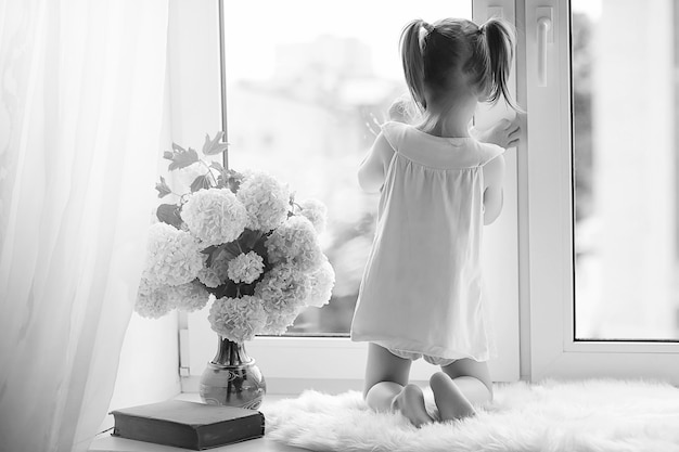 Auf der Fensterbank sitzt ein kleines Mädchen. Ein Blumenstrauß in einer Vase am Fenster und ein Mädchen, das Blumen schnüffelt. Eine kleine Prinzessin in einem weißen Kleid mit einem Strauß weißer Blumen am Fenster.