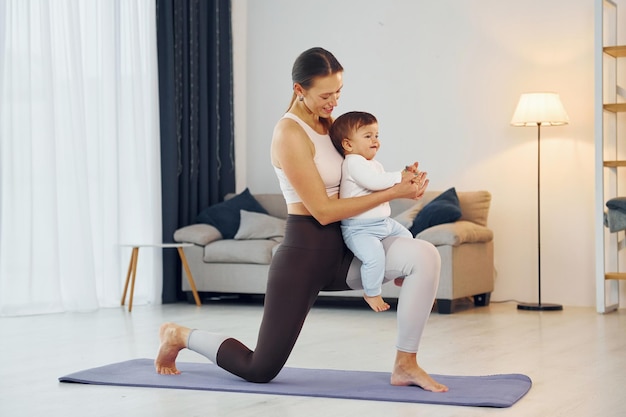 Auf der Matte stehen und Übungen machen Mutter mit ihrer kleinen Tochter ist zusammen zu Hause