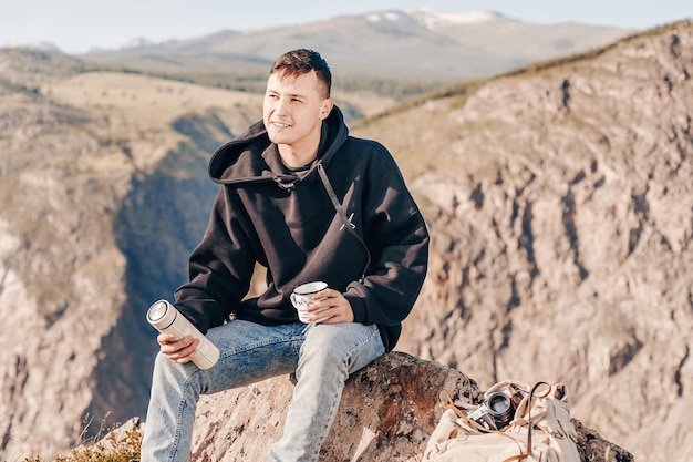 Auf der Wanderung ein heißes Getränk trinken. Junger Wanderer in schwarzem Hoodie sitzt auf Felsen und hält Thermoskanne und weißen Reisebecher in den Händen. Ein Blick zur Seite und ein Lächeln. Hintergrund von felsigen Hügeln.