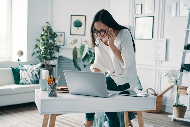 Auf die Anforderungen der Kunden hören. Schöne junge Frau, die Laptop benutzt und telefoniert, während sie im Home Office arbeitet