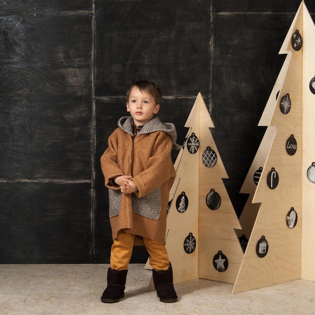 Auf dunklem Hintergrund posiert kleiner Junge in einem stilvollen Mantel in der Nähe eines Weihnachtsbaums aus Holz