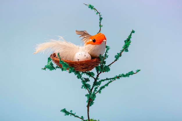 Auf einem dekorativen Baum ein Nest mit Eiern und ein kleiner gelber Vogel auf blauem Hintergrund, Nahaufnahme mit Kopierraum