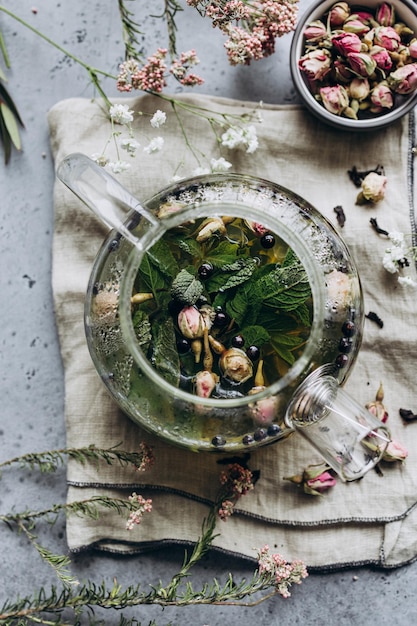 Auf einem grauen Hintergrund von oben eine offene Teekanne mit Kräutertee und getrockneten Rosenblüten