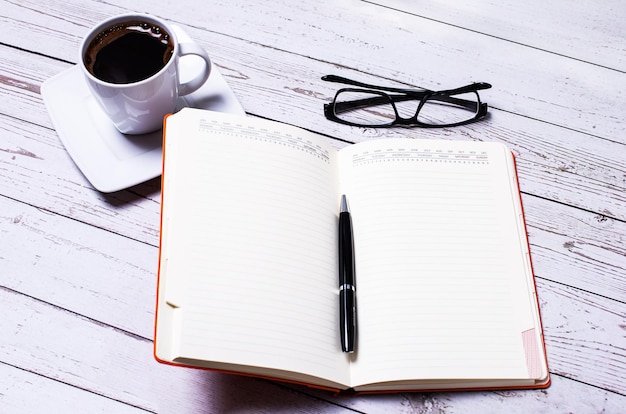 Auf einem hellen Holztisch befindet sich ein offenes Notizbuch mit einem Stift, einer weißen Tasse Kaffee und Gläsern Geschäftskonzept Arbeitsplatz aus nächster Nähe