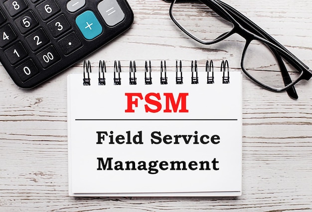 Auf einem hellen Holztisch Rechnerbrille und ein leerer Notizblock mit dem Text FSM Field Service Management Business Concept