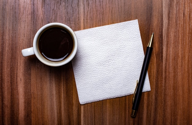 Auf einem Holztisch neben einer weißen Tasse Kaffee und einem Stift steht eine saubere weiße Papierserviette. Speicherplatz kopieren