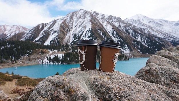 Auf einem Stein stehen zwei Pappbecher für Kaffee. Auf dem Hintergrund eines Bergsees