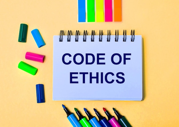 Auf einer beigen Oberfläche ein Notizbuch mit den Worten CODE OF ETHICS, helle Filzstifte und Aufkleber