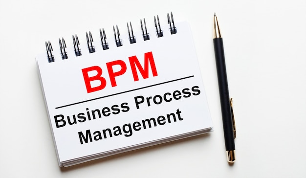 Auf einer hellen Oberfläche befinden sich ein weißes Notizbuch mit den Worten BPM Business Process Management und ein Stift.
