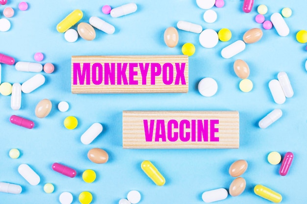 Auf hellblauem Hintergrund bunte Pillen und Holzklötze mit dem Text MONKEYPOX VACCINE Pharmaceutics Medical Concept
