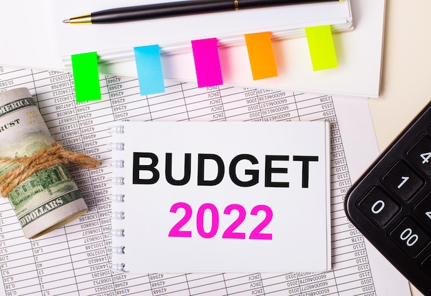 Auf hellem Hintergrund ein Stift, Dollar, ein Notizbuch mit dem Text BUDGET 2022 und helle Aufkleber. Geschäftskonzept