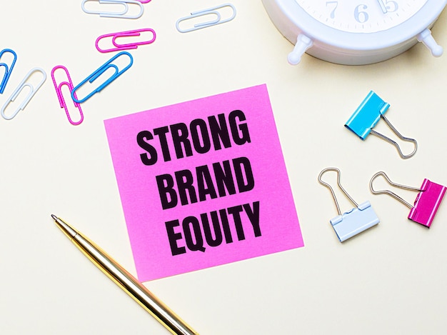 Auf hellem Hintergrund ein weißer Wecker, pinke, blaue und weiße Büroklammern, ein goldener Stift und ein pinker Sticker mit dem Text STRONG BRAND EQUITY