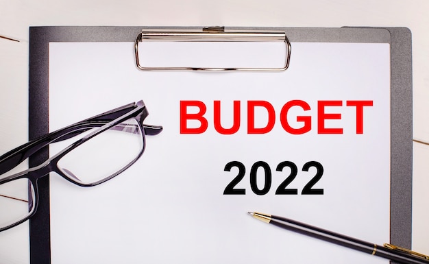 Auf hellem Holzhintergrund eine Brille, ein Stift und ein Blatt Papier mit dem Text BUDGET 2022. Geschäftskonzept