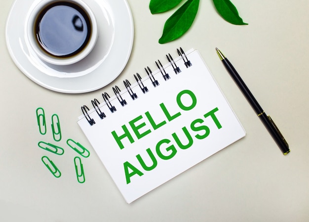 Auf hellgrauem Hintergrund eine weiße Tasse Kaffee, grüne Büroklammern und ein grünes Blatt einer Pflanze sowie ein Stift und ein Notizbuch mit den Worten HALLO AUGUST.