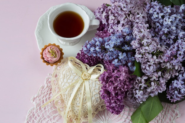 auf rosa hintergrund ein fliederstrauss, eine tasse tee, ein kuchen und ein herz aus spitze