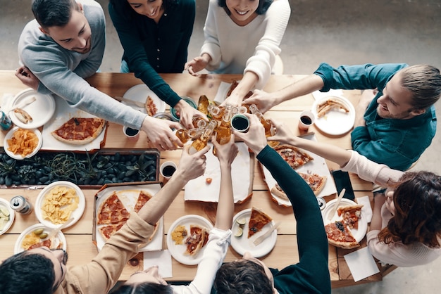 Auf unsere Freundschaft! Draufsicht auf junge Leute in Freizeitkleidung, die sich gegenseitig anstoßen und lächeln, während sie eine Dinnerparty im Haus veranstalten