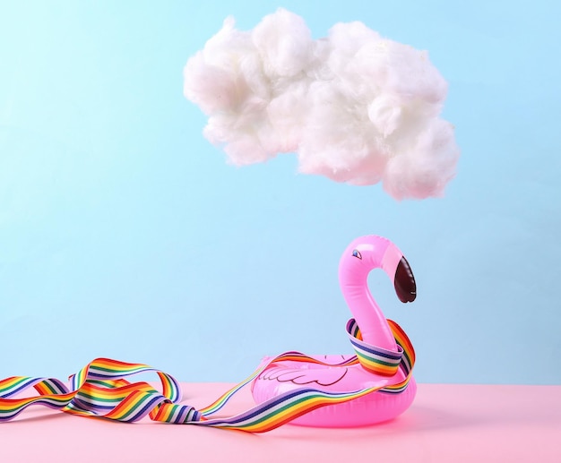 Aufblasbarer Flamingo, eingewickelt in Regenbogenband und schwebende, flauschige Wolke auf einem blaurosa Pastellhintergrund Konzeptkunst Minimalistische Idee