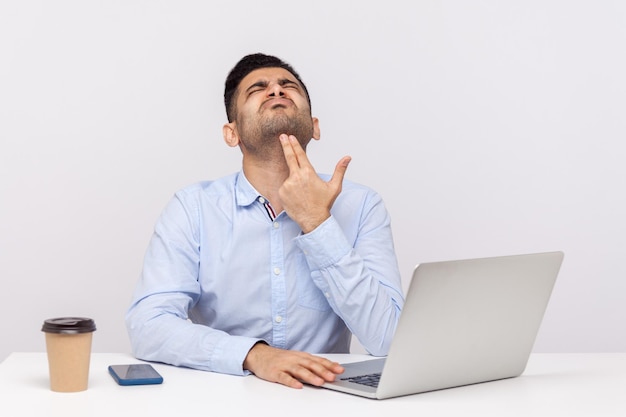 Aufgebrachter, depressiver männlicher Angestellter sitzt im Büro mit Laptop auf dem Schreibtisch und zeigt mit dem Finger auf den Kopf