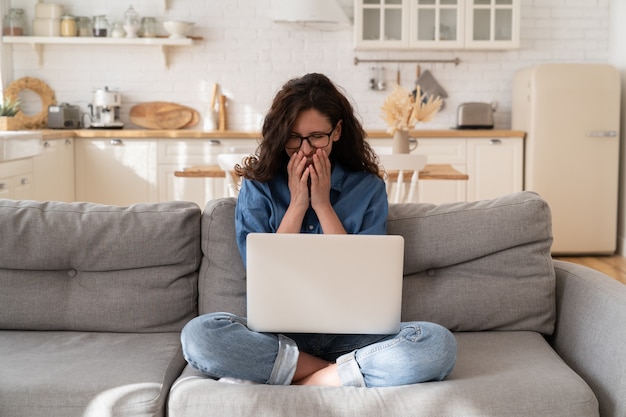 Aufgeregt fröhliche Frau mit Brille, die lächelnd E-Mails auf dem Laptop mit guten Nachrichten liest oder lustige Videos ansieht