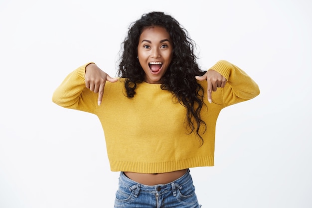 Aufgeregt glückliche junge afroamerikanische süße Frau in gelbem Pullover, die erstaunt nach unten keucht, coole Werbung fördert, Kamera anstarrt, tolles Angebot über weiße Wand vorschlägt