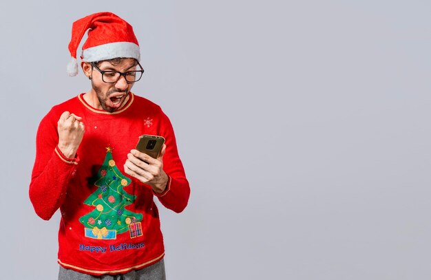 Aufgeregt Kerl in Weihnachtsmütze Blick auf Handy und Feiern Menschen in Weihnachtsmütze Blick auf Handy aufgeregt Aufgeregter junger Mann in Weihnachtsmütze mit Handy feiern