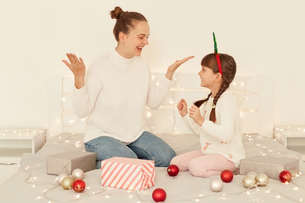 Aufgeregt lächelnde glückliche Mutter und Tochter, die weiße Pullover im lässigen Stil tragen, die auf dem Bett mit Weihnachtsdekorationen sitzen, glücklich schreien und das neue Jahr feiern.