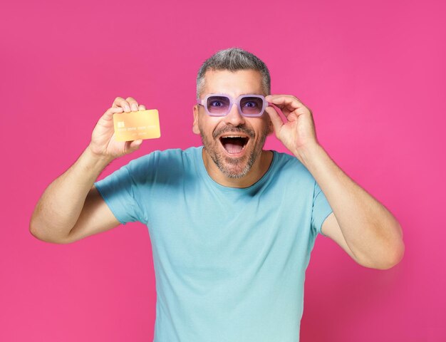 Aufgeregt, schockiert, gutaussehender Mann mittleren Alters hält Debit-Kreditkarte in der Hand und repariert seine Brille. Er trägt ein blaues T-Shirt auf rosa Hintergrund. Finanzbanking-Konzept