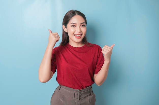 Aufgeregte asiatische Frau, die ein rotes T-Shirt trägt, gibt Daumen nach oben Handgeste der Zustimmung, isoliert durch blauen Hintergrund