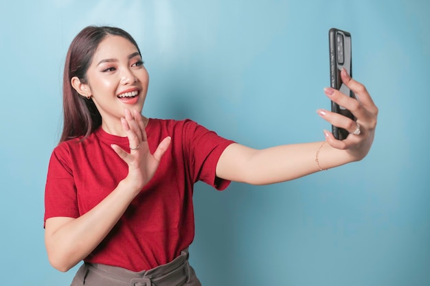 Aufgeregte asiatische Frau, die ein rotes T-Shirt trägt und auf ihr Smartphone zeigt, isoliert durch einen blauen Hintergrund