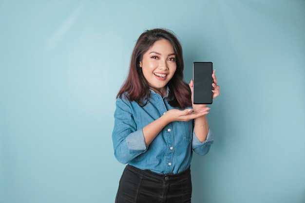 Aufgeregte asiatische Frau mit blauem Hemd, die auf den Kopierbereich auf ihrem Smartphone zeigt, isoliert durch blauen Hintergrund