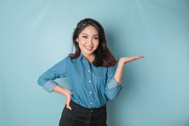 Aufgeregte asiatische Frau mit blauem Hemd, die auf den Kopierbereich neben ihr zeigt, isoliert durch blauen Hintergrund