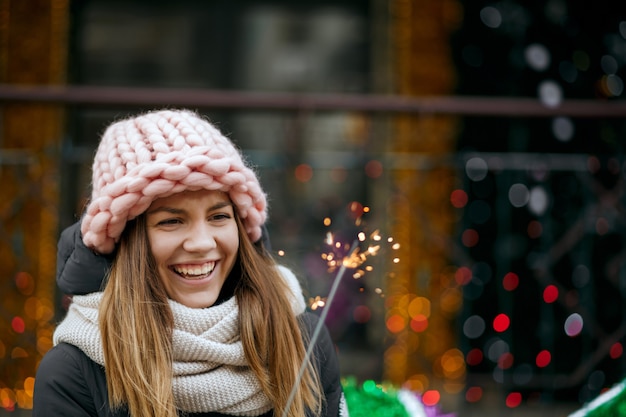 Aufgeregte blonde Frau mit Winteroutfit feiert Neujahr auf der Straße mit Wunderkerzen. Platz für Text