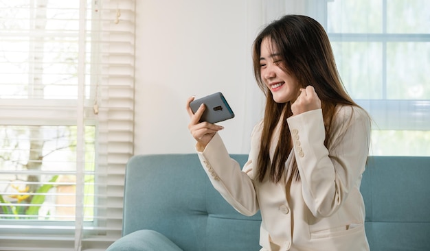 Aufgeregte Frau benutzt ein Handy, das die Faust hochhebt, sie feiert den Sieg online oder erhält gute Nachrichten auf dem Sofa im Wohnzimmer zu Hause, glückliche asiatische Frau lustig auf dem Smartphone, die sich überrascht und aufgeregt fühlt