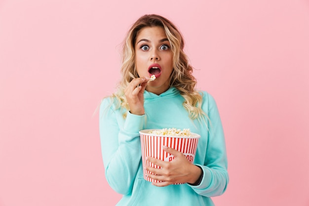 Aufgeregte Frau, die Eimer mit Popcorn hält und Kamera betrachtet, lokalisiert über rosa Wand